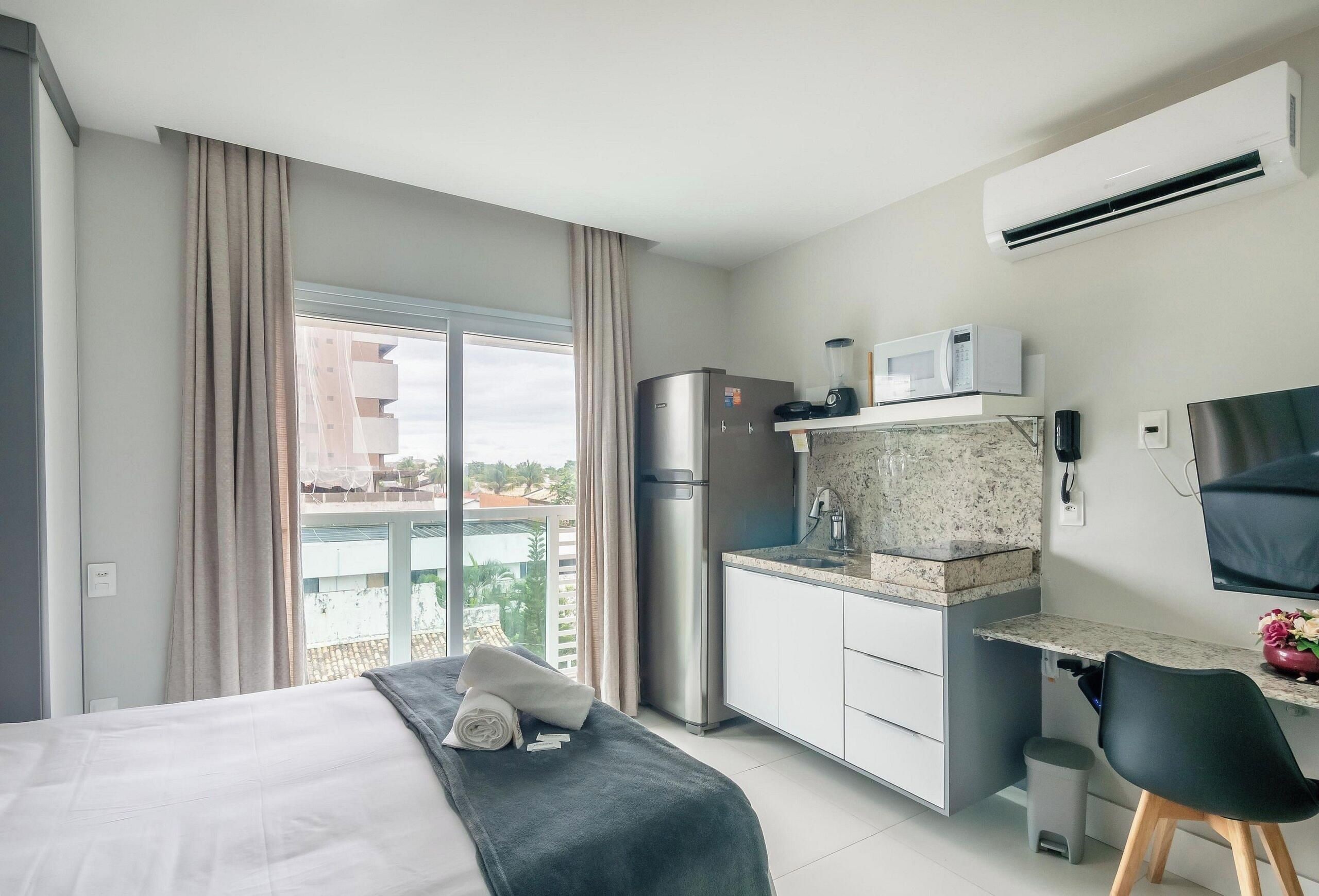 A modern hotel room with a bed, a kitchenette, a large window with curtains, and a view of buildings outside.