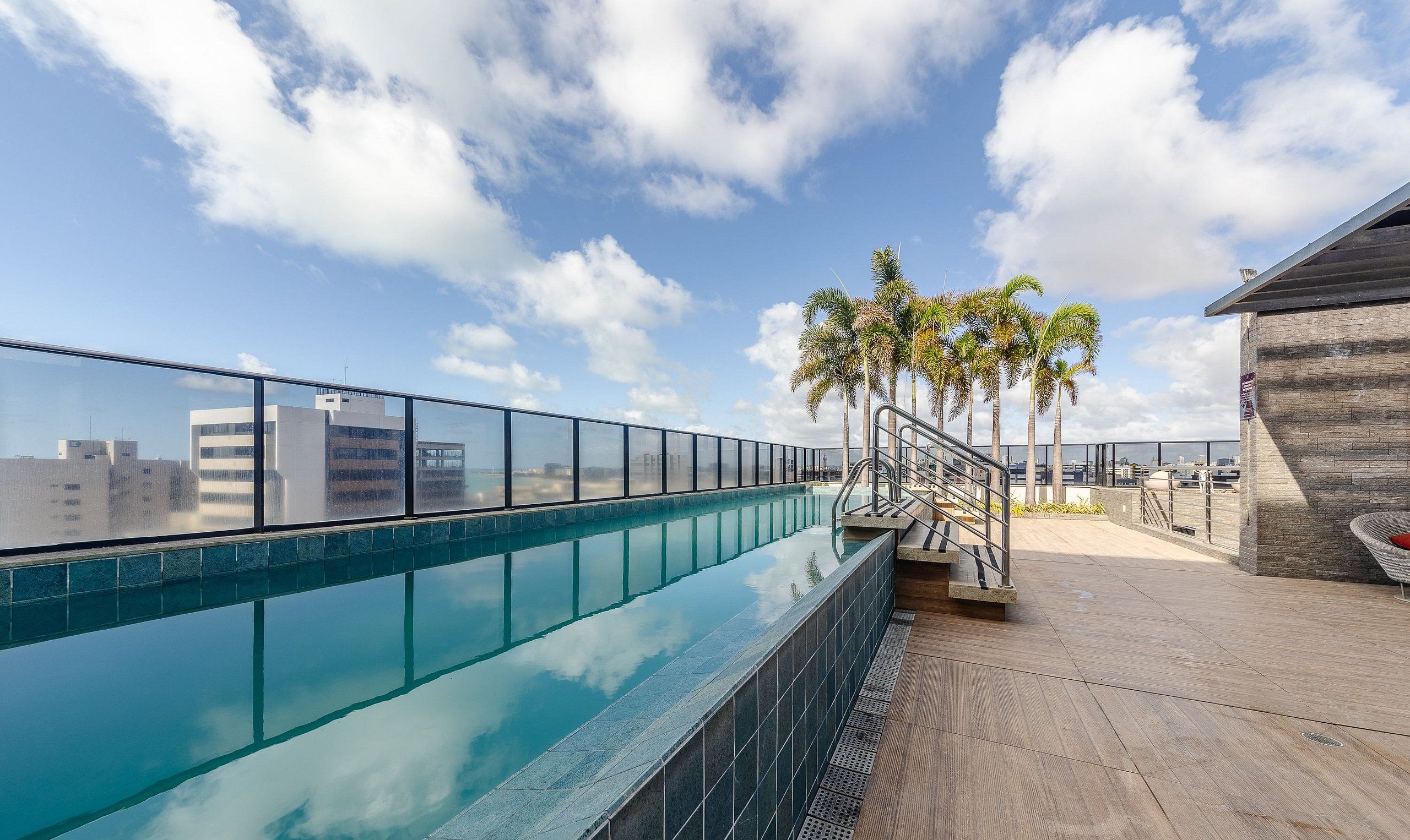 Una piscina en la azotea con agua azul cristalina, barandilla de vidrio, palmeras y vista panorámica de la ciudad.