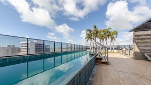 Una piscina al terrat amb aigua blava cristal·lina, barana de vidre, palmeres i vista a la ciutat.