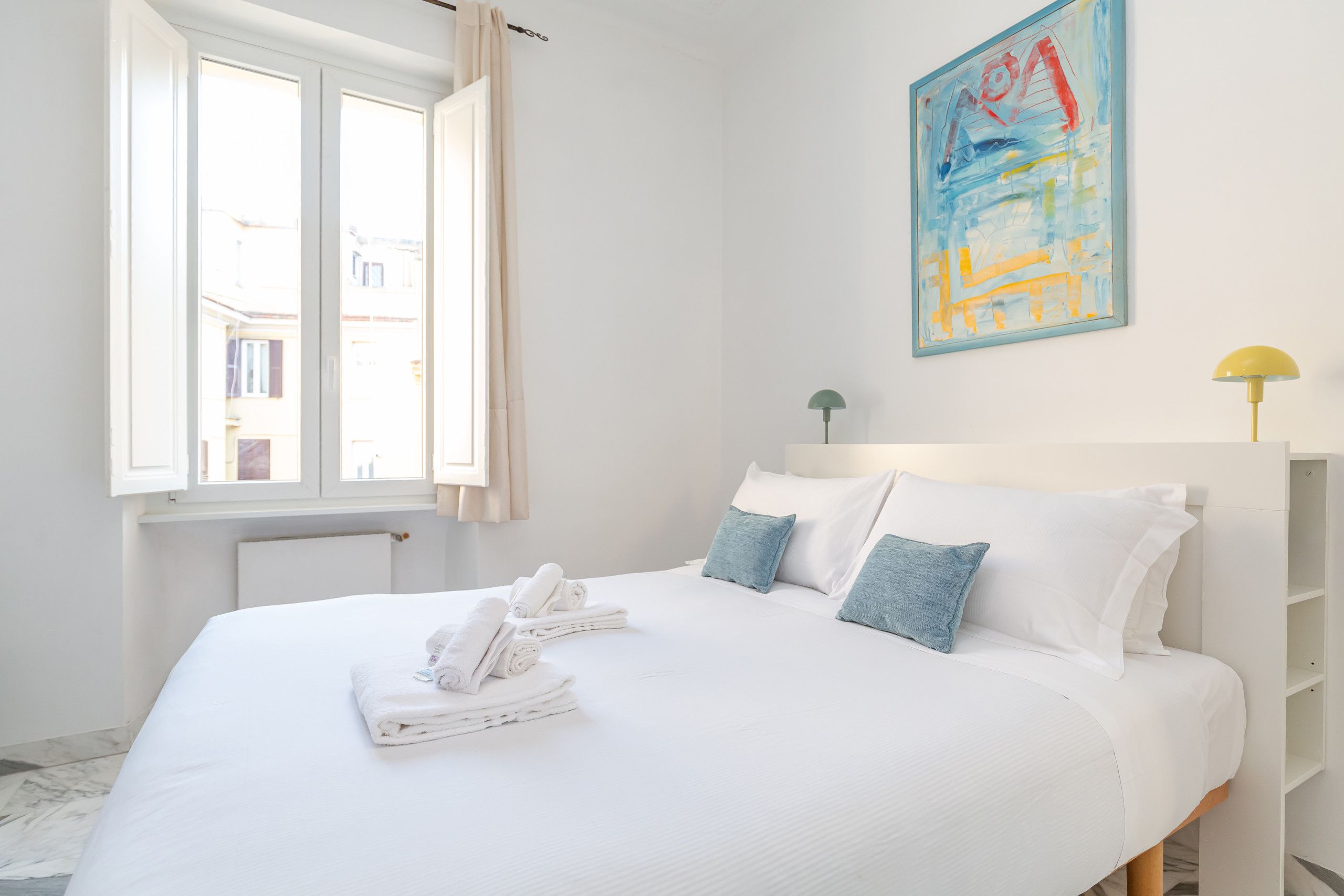 A hotel room with a bed, pillows, a painting, a lamp, and a window.