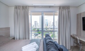 Ein Hotelzimmer mit Bett, Sofa, Fernseher und Blick auf eine Stadtlandschaft durch große Fenster.