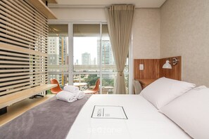 A hotel room with a large window, a bed, a wooden headboard, a chair, and a small table. - Flat in Full Condo, Setor Bueno - Txp0903 (Goiânia)