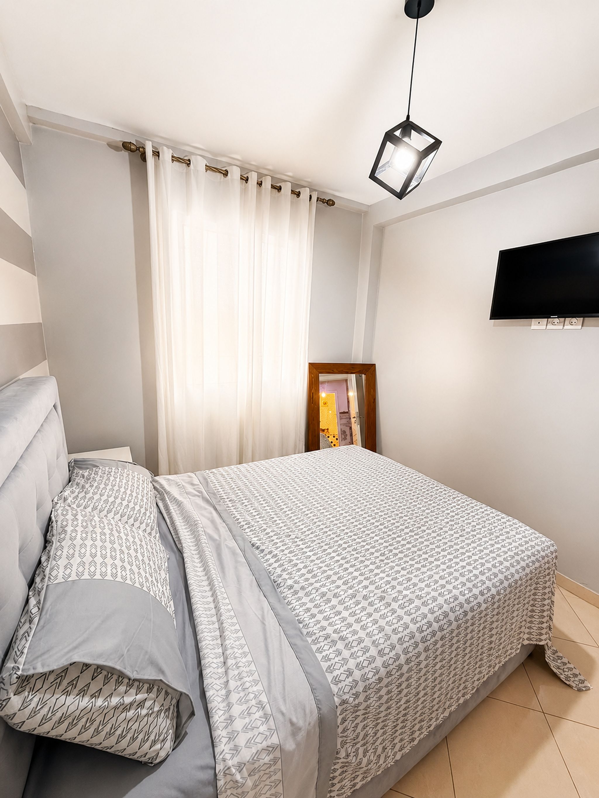 Un dormitorio con cama, televisor montado en la pared y ventana con cortinas.