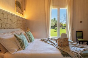 Une chambre à coucher avec un lit, des oreillers, un panier en osier, une chaise et une vue sur un jardin par la fenêtre.