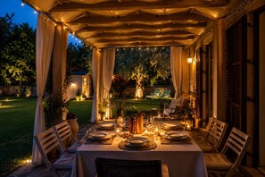 An outdoor dining area with a covered table set for four, surrounded by chairs, and a view of a garden and house in the background. - Villa Ulìa Sicilymyhome (Ispica)