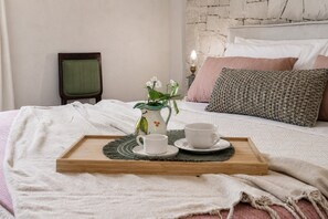 Ein ordentlich bezogenes Bett mit einer Tablett voller weißer Tassen und einer Blumenvase, ein Nachttisch mit einer Lampe sowie eine Steinsäule im Hintergrund.