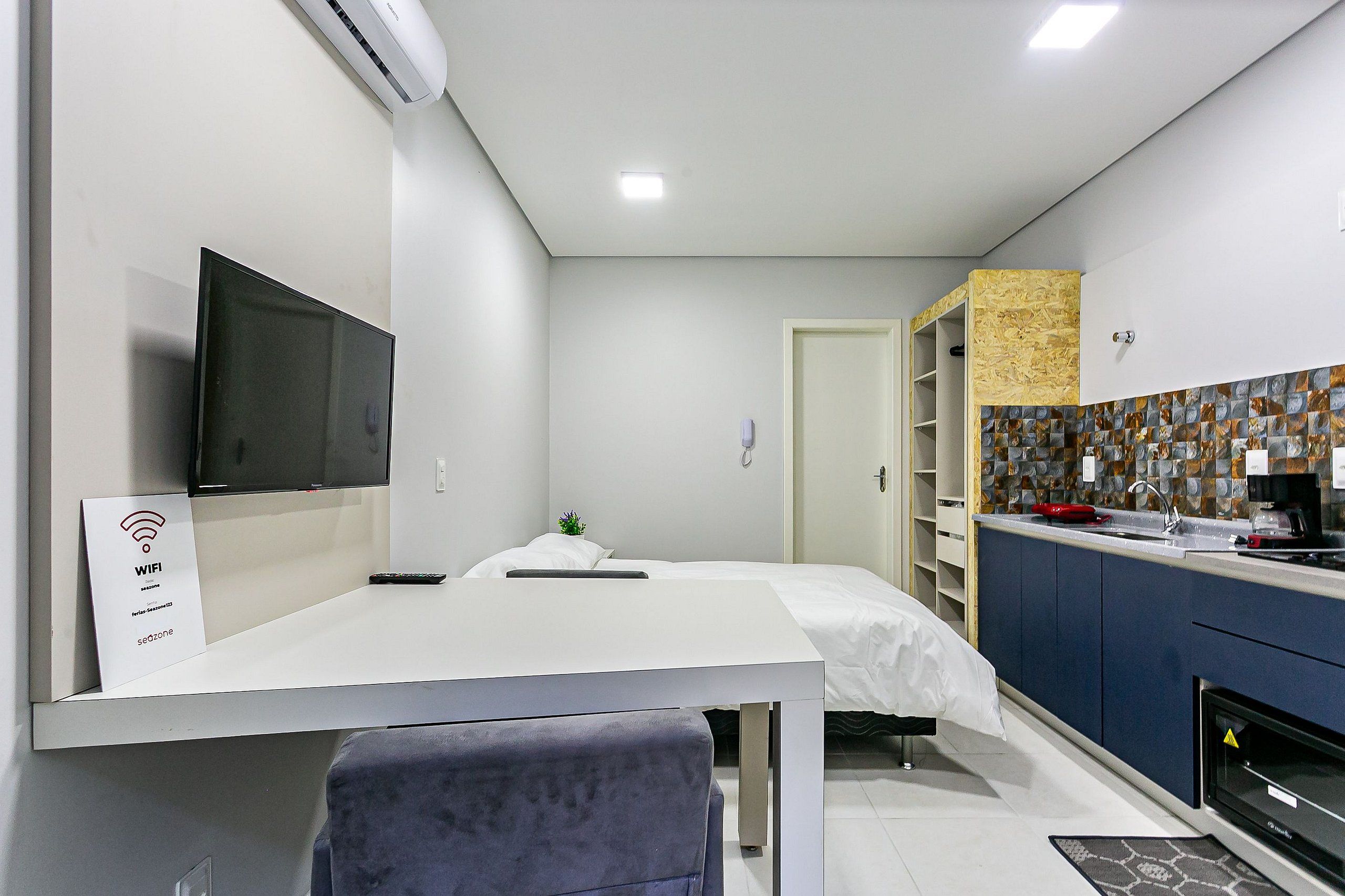 Una habitación de hotel moderna con televisión de pantalla plana, una cama con ropa de cama blanca, una mesita de noche pequeña, un escritorio con una silla, una kitchenette con estufa y un baño con ducha.