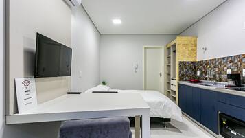 Una habitación de hotel moderna con televisión de pantalla plana, una cama con ropa de cama blanca, una mesita de noche pequeña, un escritorio con una silla, una kitchenette con estufa y un baño con ducha.