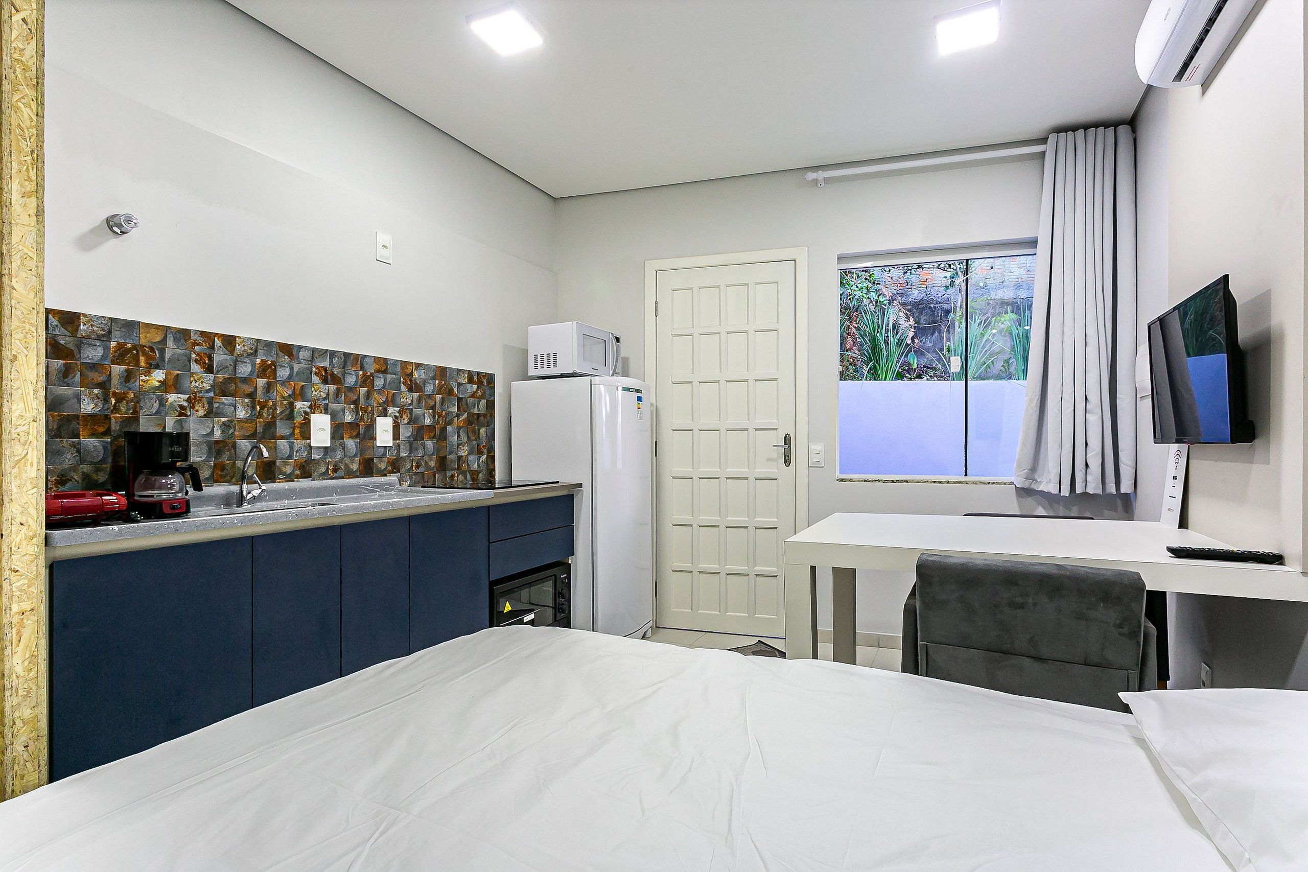 Habitación de hotel compacta con kitchenette, una cama, un escritorio con silla, televisión y ventana con cortinas.