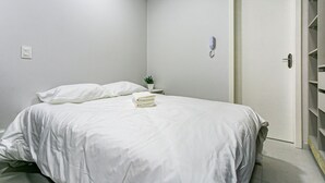 A neatly made bed with white bedding, a bedside table with a plant, and a closed door. - Cozy Studio Facing the Lagoon Sdr007 (Florianópolis)