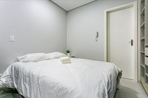 Een opgemaakt bed met witte lakens, een nachtkastje met een plantje, en een gesloten deur.