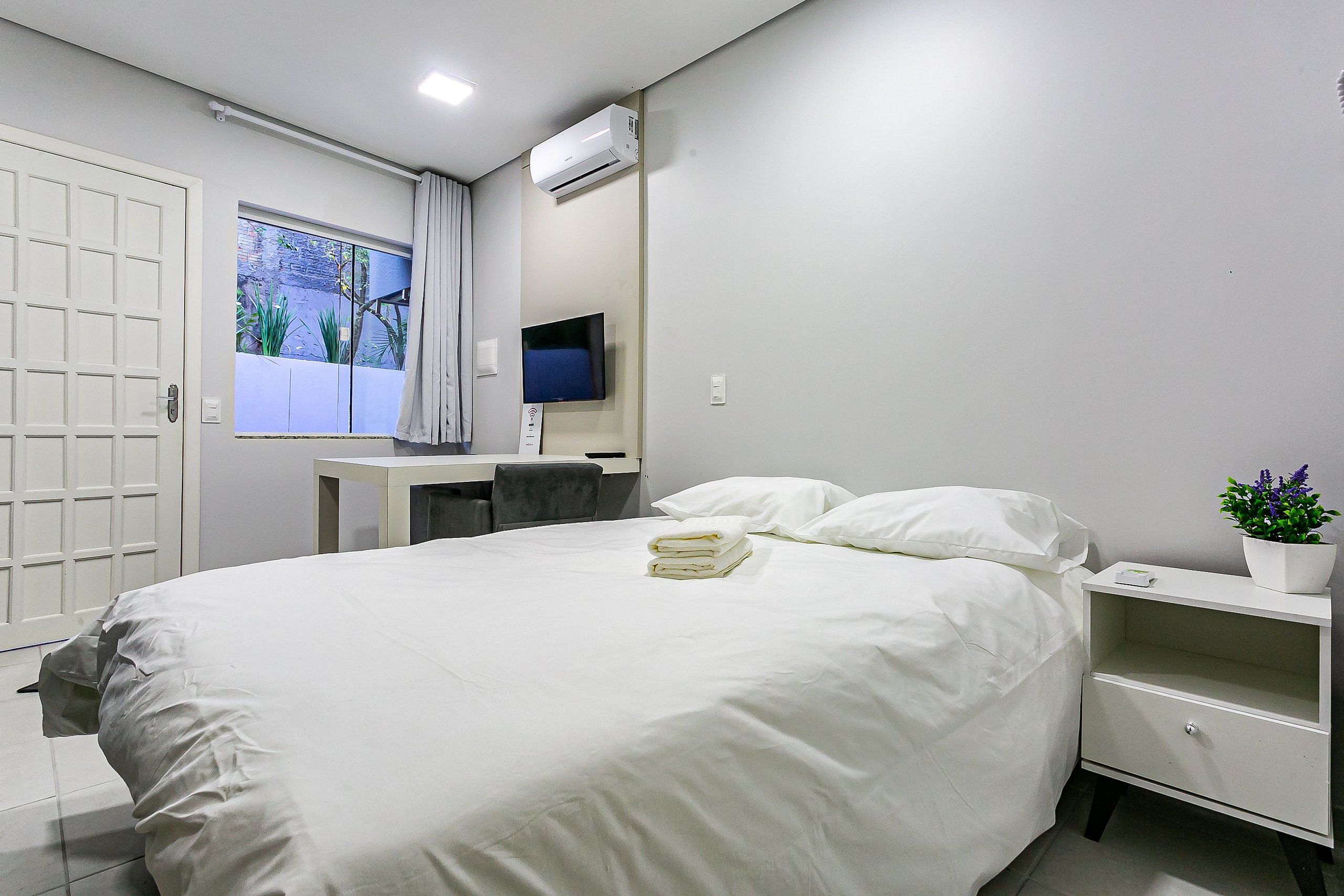 Una cama bien hecha con ropa blanca, una mesita de noche con una planta y un televisor montado en la pared.