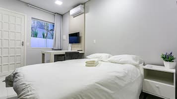 Una cama bien hecha con ropa blanca, una mesita de noche con una planta y un televisor montado en la pared.