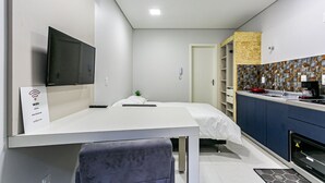 A modern hotel room with a flat-screen TV, a bed with white bedding, a small bedside table, a desk with a chair, a kitchenette with a stove, and a bathroom with a shower. - Cozy Studio Facing the Lagoon Sdr007 (Florianópolis)