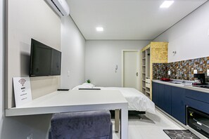 Een moderne hotelkamer met een flatscreen-televisie, een bed met wit beddengoed, een klein nachtkastje, een bureau met stoel, een kitchenette met kookplaat en een badkamer met douche.