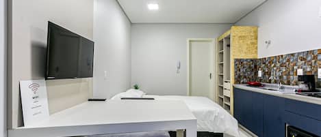 A modern hotel room with a flat-screen TV, a bed with a white mattress, a small bedside table, a chair, a kitchenette with a microwave, and a bathroom with a shower.