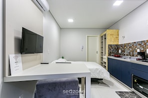 Quarto de hotel moderno com televisão de tela plana, cama com colchão branco, pequena mesa de cabeceira, uma cadeira, cozinha compacta com micro-ondas e banheiro com chuveiro.