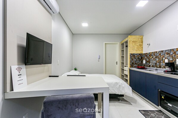 Habitación de hotel moderna con televisor de pantalla plana, cama con colchón blanco, mesita de noche pequeña, una silla, kitchenette con microondas y baño con ducha.