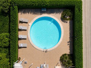 Eine Vogelperspektive auf einen runden Swimmingpool, umgeben von einer gefliesten Liegefläche mit Liegestühlen und einem kleinen Gartenbereich.