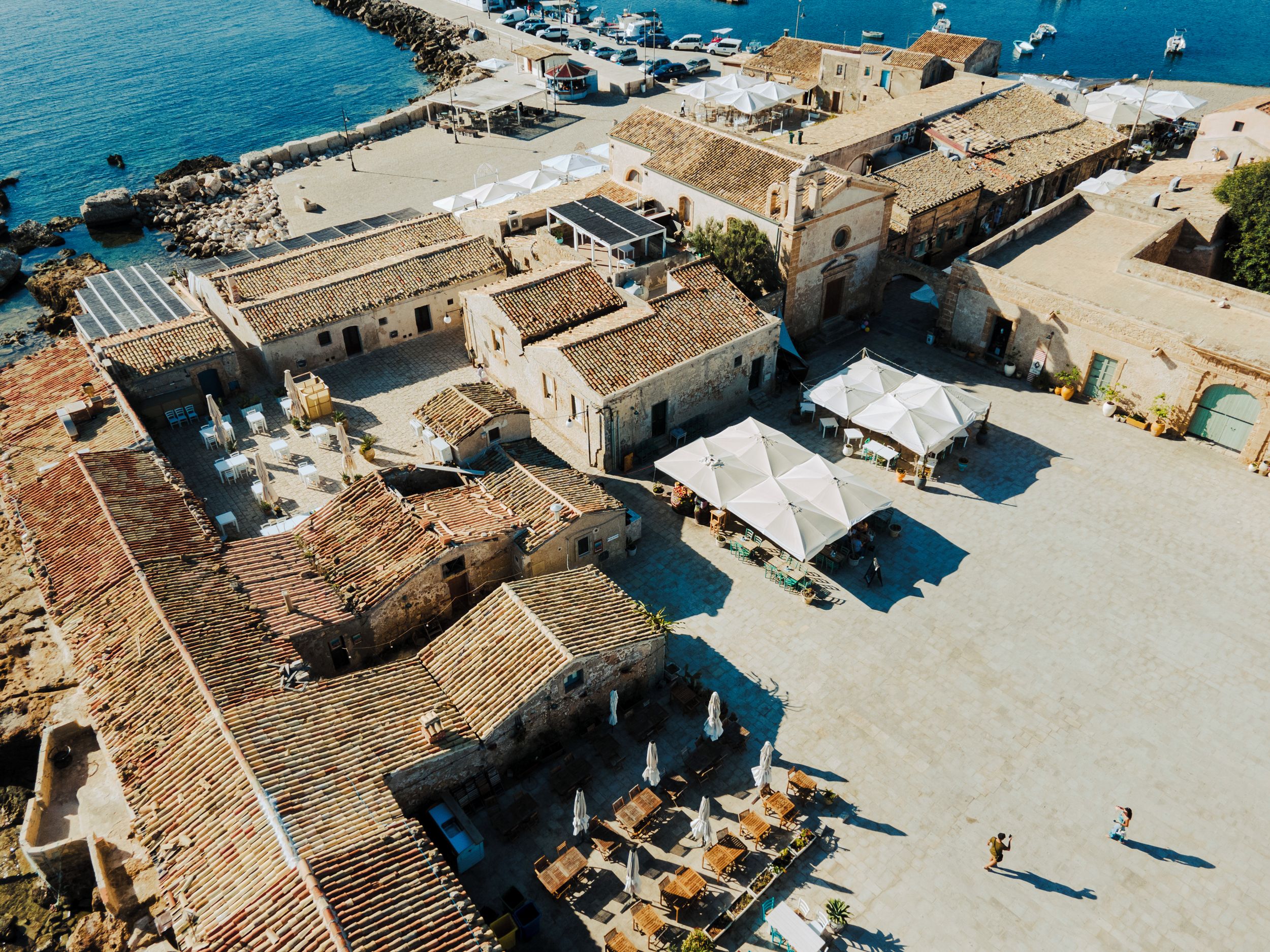 Vue aérienne d’une ville côtière avec des bâtiments aux toits de tuiles rouges, des espaces de restauration en plein air et une marina où sont amarrés des bateaux.