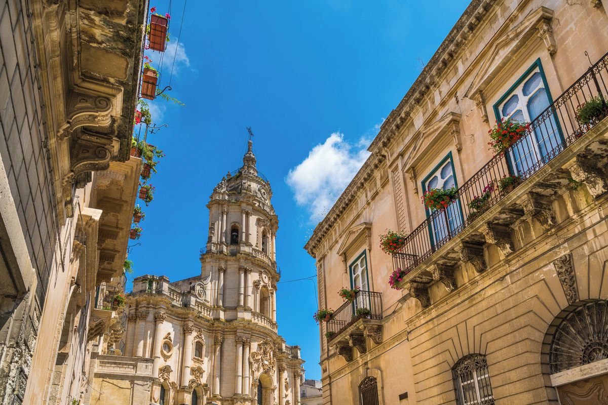 Un edificio histórico con arquitectura ornamentada, una torre de campanas y balcones con jardineras florales.