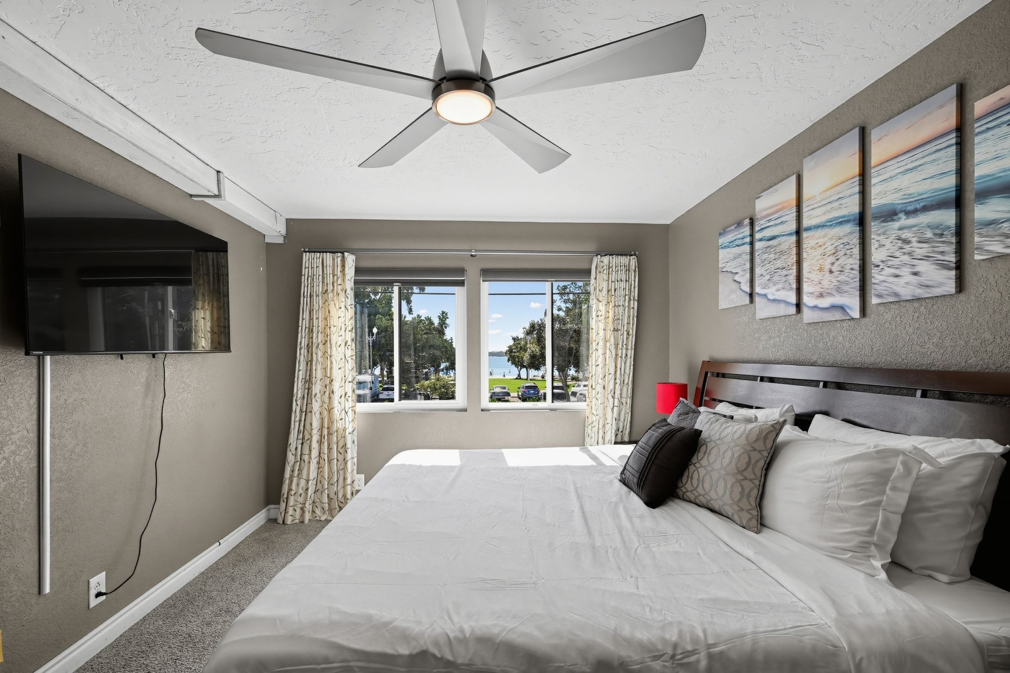 A bedroom with a bed, a ceiling fan, a television, and a view of the outdoors.