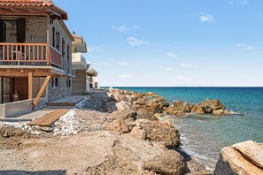 Un edifici a la costa amb façana de pedra, terrassa de fusta i vistes al mar.