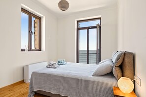 A bedroom with a bed, a bedside table with a lamp, a window, and a view of the sea.