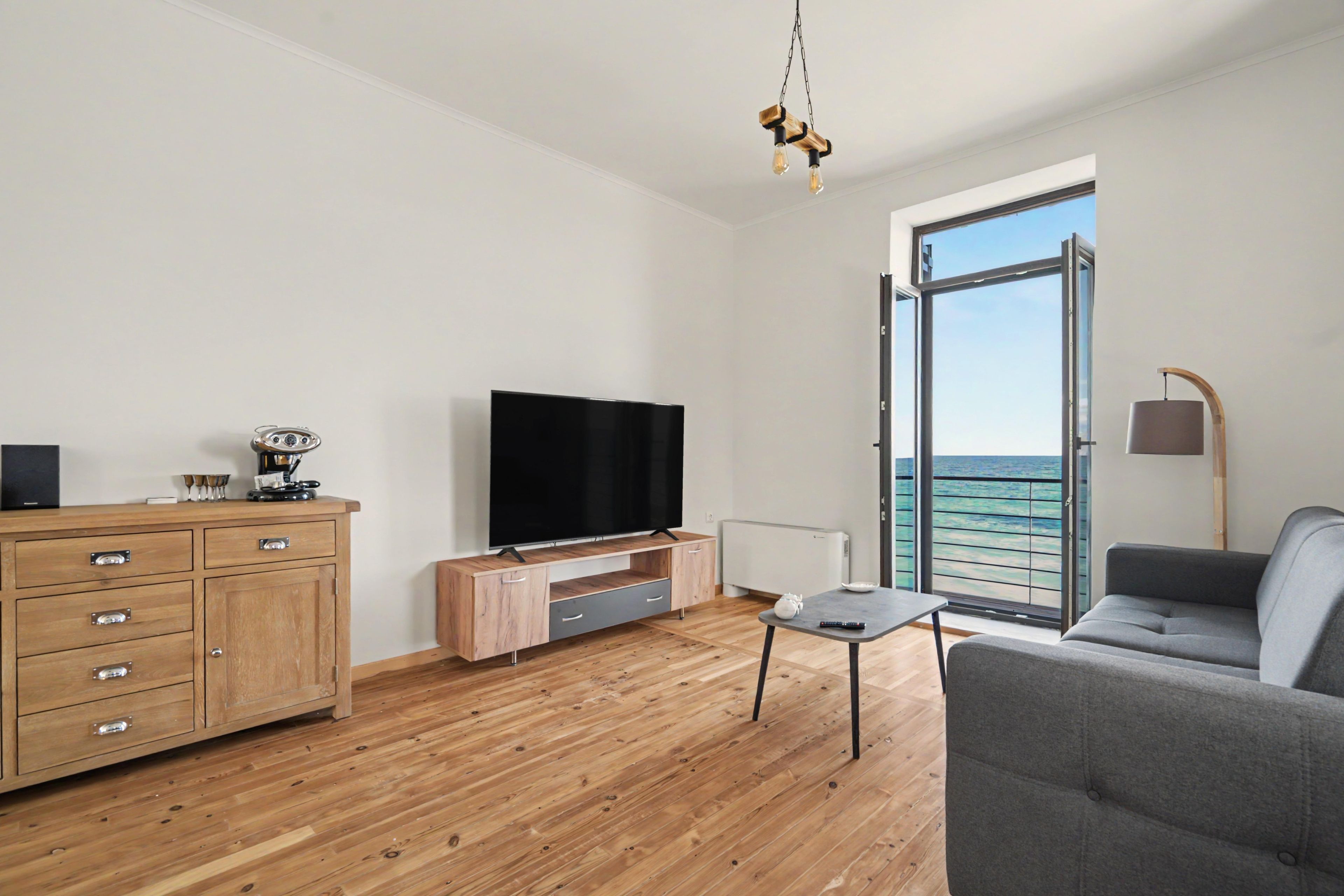 Een moderne woonkamer met een flatscreen televisie, houten meubels, een grijze bank en uitzicht op de zee door een glazen deur.
