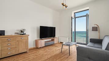 Een moderne woonkamer met een flatscreen televisie, houten meubels, een grijze bank en uitzicht op de zee door een glazen deur.