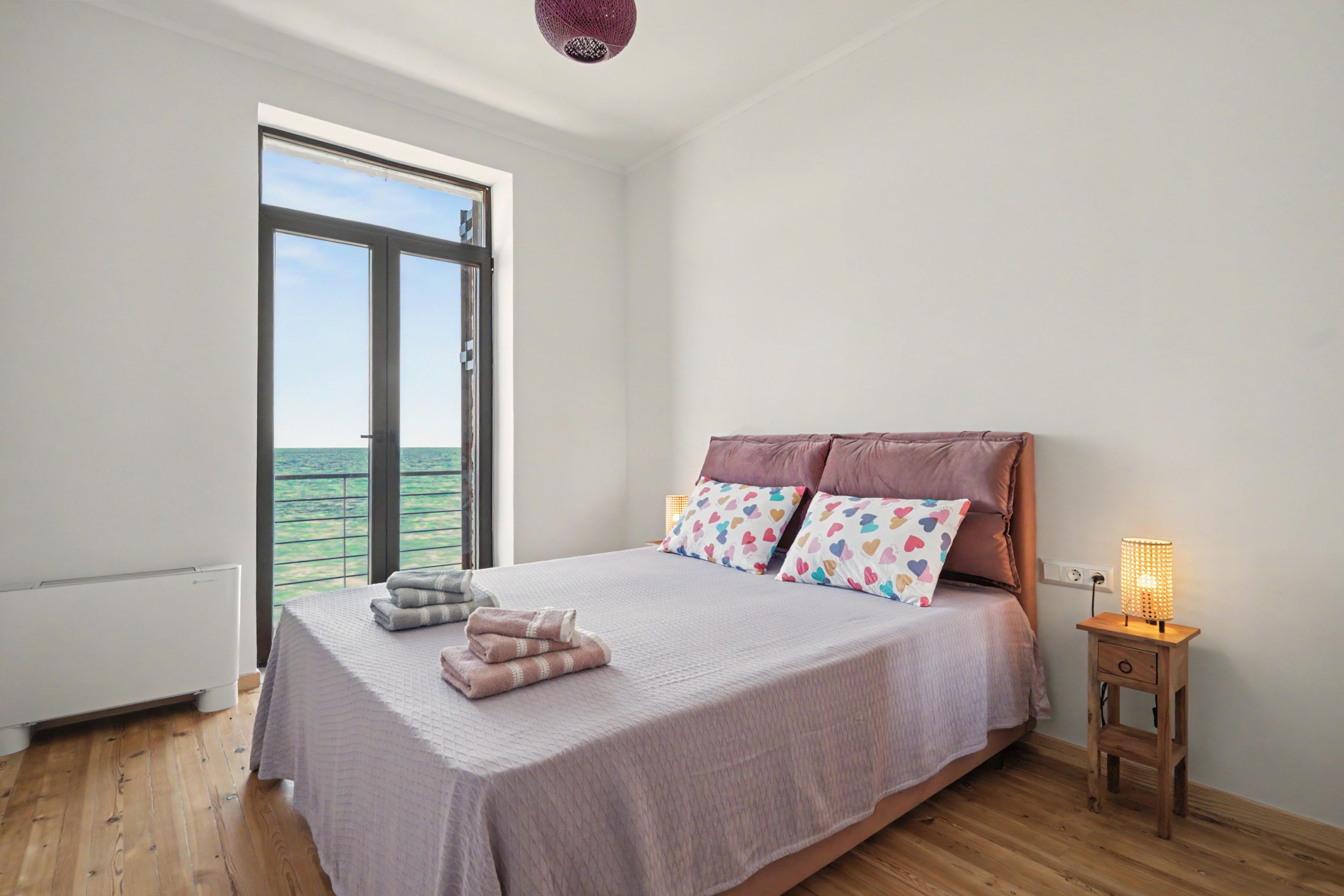 Une chambre avec une grande fenêtre, un lit avec des oreillers, une table de chevet avec une lampe et un plancher en bois.