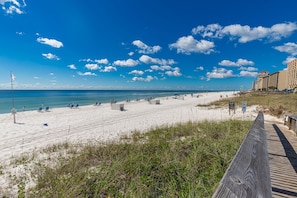 A beach with a wooden boardwalk leading to the shore, clear skies, and a distant view of a hotel. - Affordable 2-bed Condo in Orange Beach, Beach Side Many Amenities Tennis villas (Orange Beach)