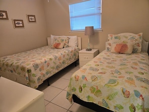 A bedroom with two beds, a window with blinds, a nightstand with a lamp, and wall art. - Affordable 2-bed Condo in Orange Beach, Beach Side Many Amenities Tennis villas (Orange Beach)