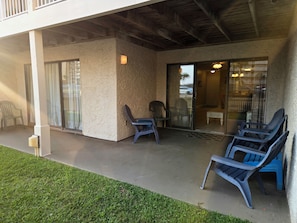A covered outdoor area with a concrete floor, two blue plastic chairs, a white table, and a glass door leading inside. - Affordable 2-bed Condo in Orange Beach, Beach Side Many Amenities Tennis villas (Orange Beach)