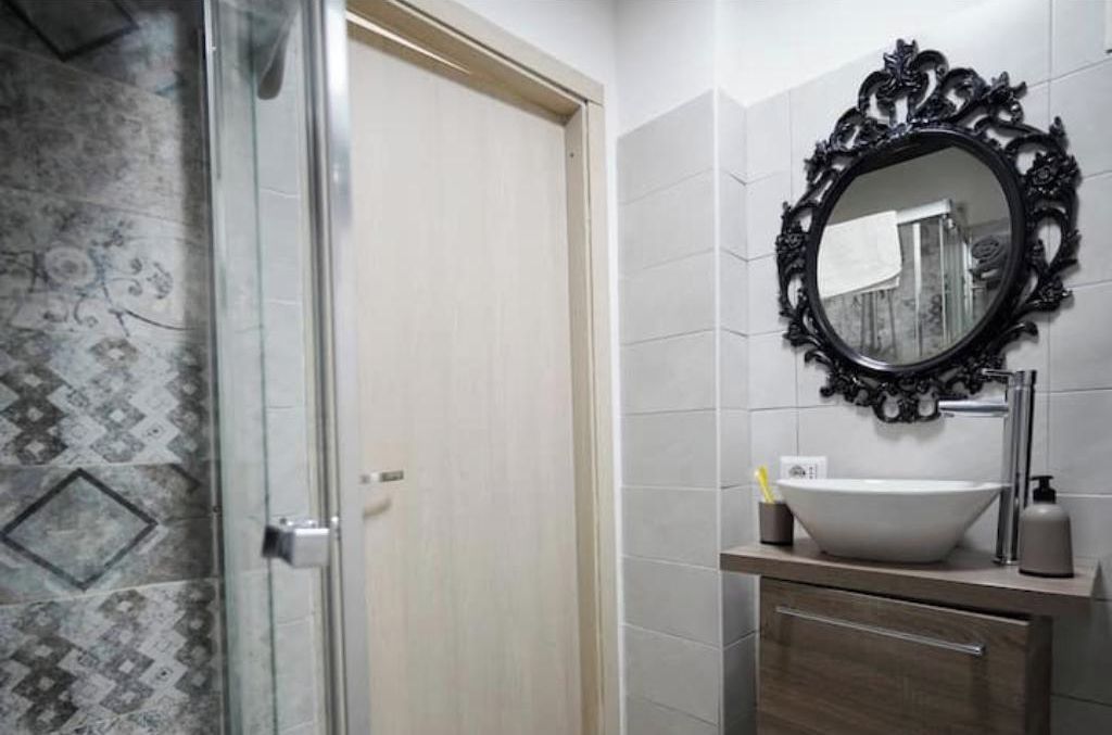 A bathroom with a glass shower enclosure, a white sink with a black ornate mirror, and patterned wall tiles.