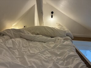A bedroom with a sloped ceiling, a bed with white bedding, a wall-mounted light fixture, and a visible power outlet. - Cozy Studio near Bastille, République & Marais (Paris)