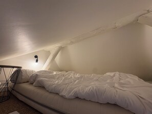 Ein Einzelbett in einem Zimmer mit schrägverdeckter Decke und einer einzelnen Lampe.