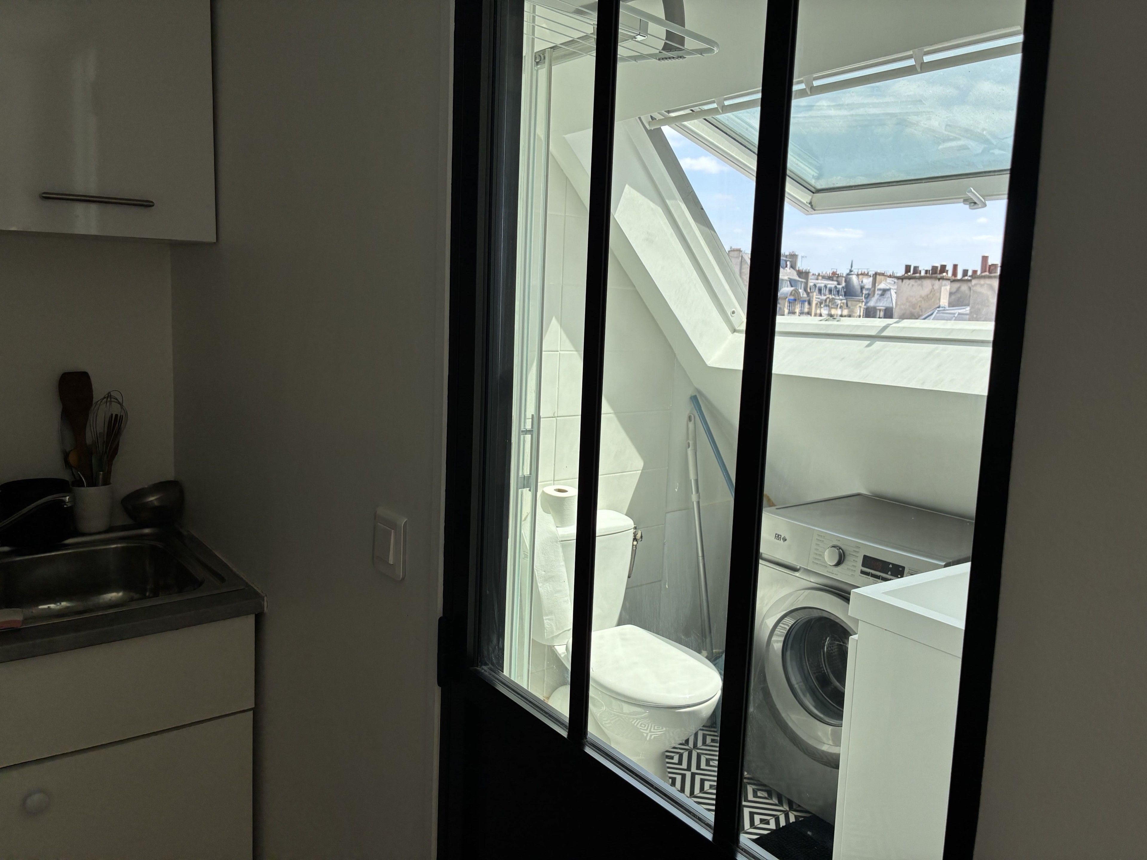Een kleine keuken met een raam, waarin een wasmachine en een toilet zichtbaar zijn.