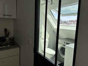 Eine kleine Küche mit Fenster, Waschmaschine und einem durch das Fenster einsehbaren WC.