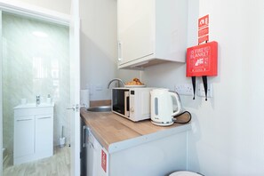 A compact kitchenette with a microwave, toaster, and kettle, a small sink, and a fire blanket mounted on the wall.