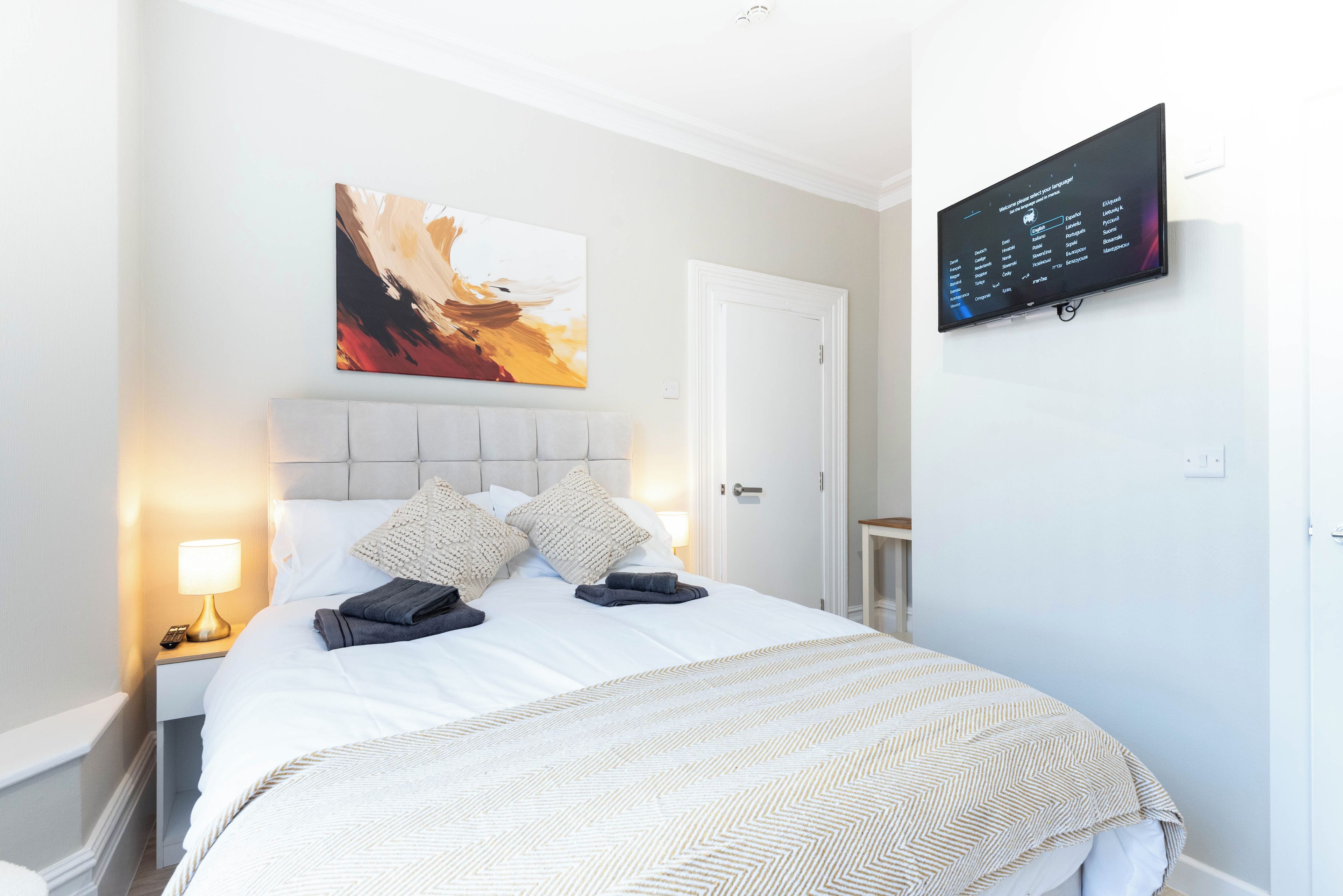 Une chambre d’hôtel avec un lit, une table de chevet, une lampe, un tableau et un écran numérique fixé au mur.