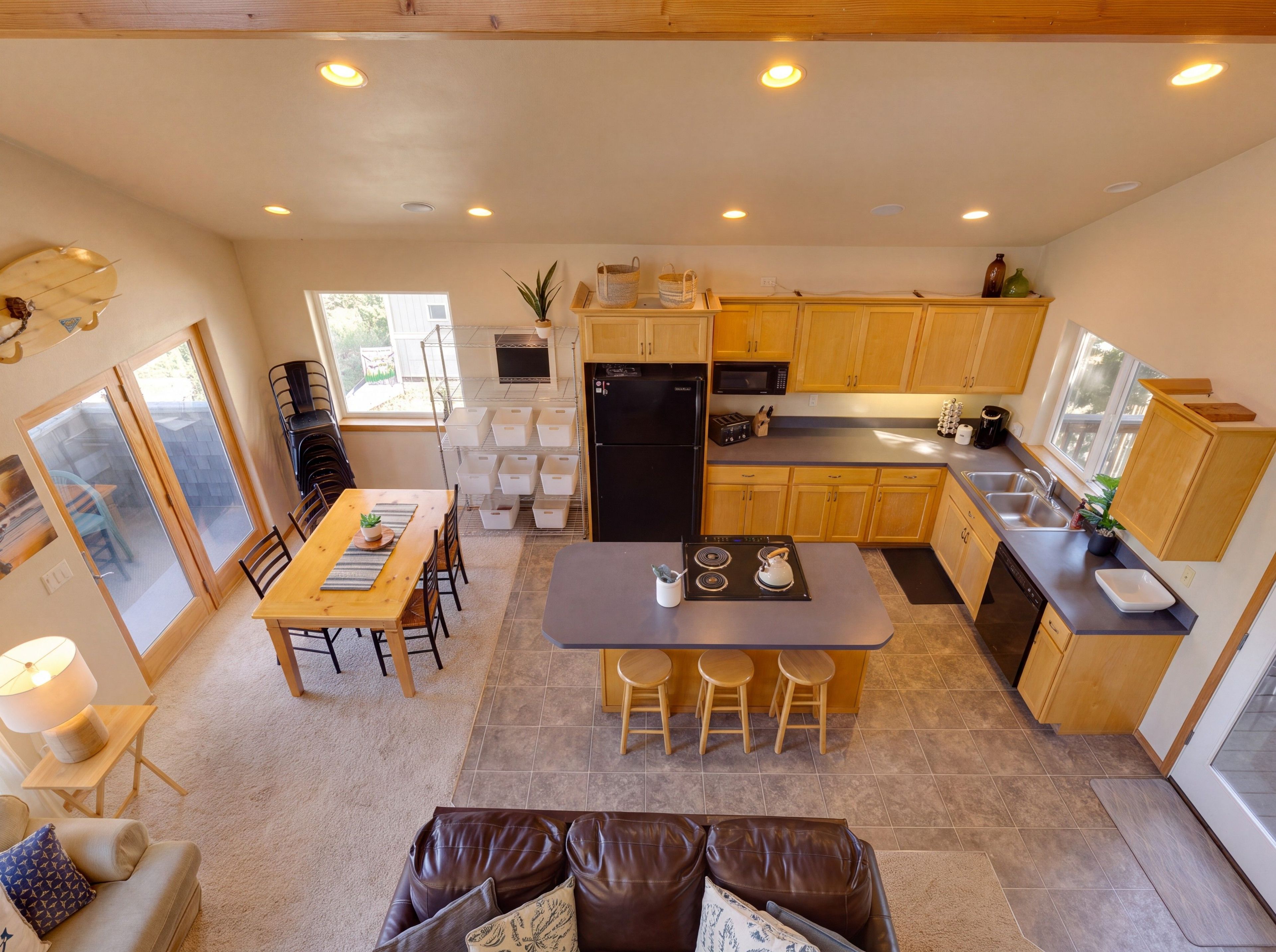 A modern kitchen with wooden cabinets, a central island with stools, a dining area with a table and chairs, and a view through a sliding glass door.
