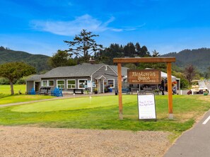 Um campo de golfe com um edifício identificado como 'Neskowin Beach Golf Course' e uma placa de sinalização com detalhes do evento.