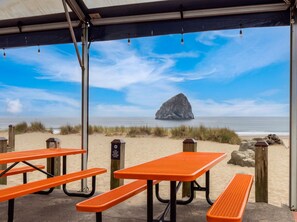 Ein Picknickbereich am Strand mit Tischen und Bänken, ein Schild, das Lärm außerhalb der Terrasse verbietet, und ein großer Felsen im Meerblick.
