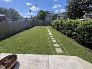 Ein Garten mit einem gepflasterten Weg, einer Holzterrasse und einem Hecken.