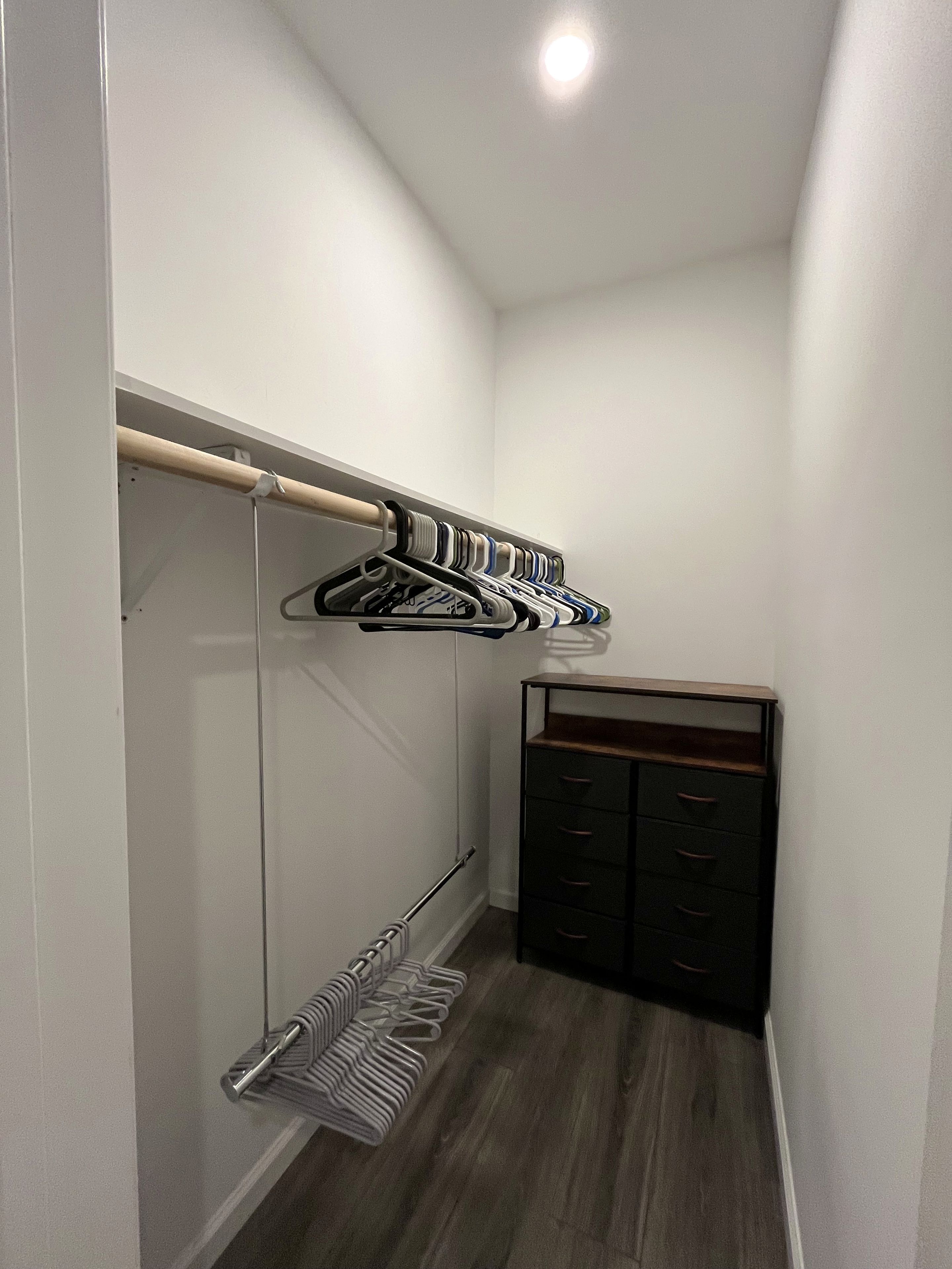 A narrow hallway with a wooden floor, a closet with dark-colored drawers, and a clothing rack with multiple hangers.