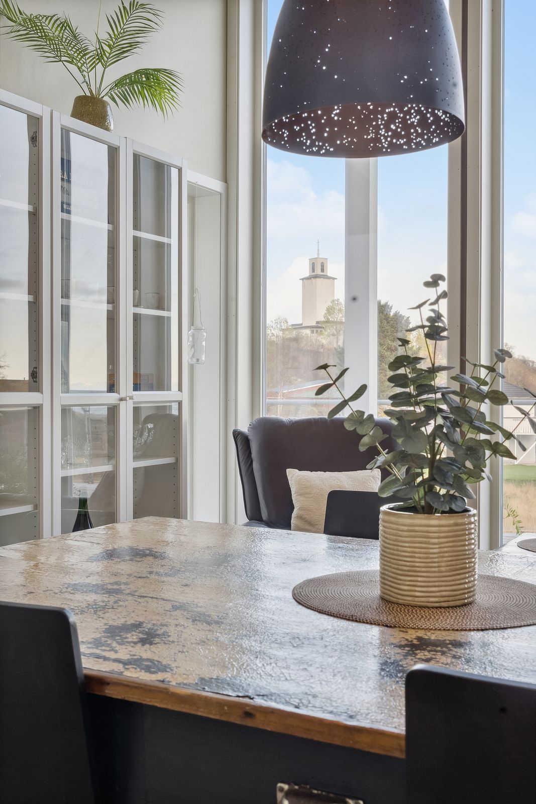 Une pièce avec une grande fenêtre, une table à manger, des chaises, une plante en pot et une lampe suspendue.