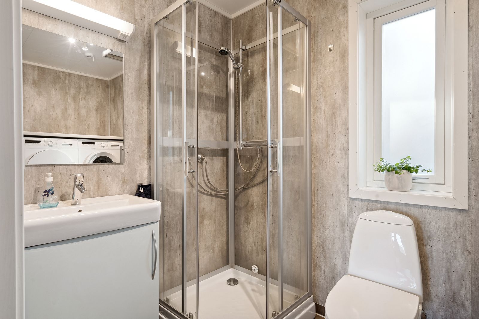 A bathroom with a glass-enclosed shower, a toilet, a sink with a mirror, and a window with a plant.