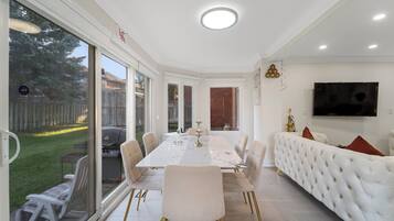 A dining area with a white table and chairs, a sofa, and a TV on the wall.