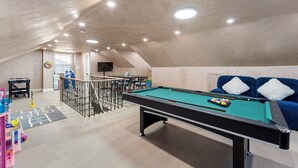 A pool table with a green felt surface, a blue sofa with white cushions, and a ping pong table in a room with a sloped ceiling and recessed lighting.
